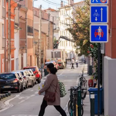 Rue avec présence d'une piétonne et d'un cycliste accompagnés de panneaux indiquant la présence d'une vélorue